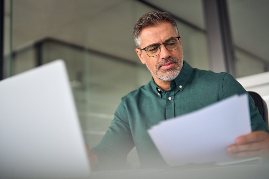 Older middle aged busy professional business man executive manager or entrepreneur working on laptop computer in office checking legal corporate paperwork, reviewing financial document management.